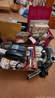 Wide view of assortment of office supplies in boxes and loose on floor. Includes phones, folders, stationery, decorative boxes, various cables, and accessories.