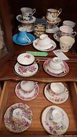 Wooden cabinet drawer with a variety of teacups and saucers including floral designs and a blue set, some stacked. Also two small blue and white ceramic napkin rings behind the teacups.