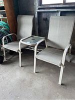 Two beige mesh patio chairs with metal frames attached on either side of a small glass top center table with umbrella hole, placed against a wall in a garage area.
