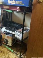 Black metal wire shelving unit with three shelves and caster wheels. Various boxed items including a trivia board game and electronics are placed on the shelves but not included.