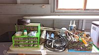 Table with a green small parts organizer with multiple drawers filled with screws, nails, nuts, bolts. Next to it is a metal tote filled with caulking tubes, hand tools like screwdrivers, pliers, and grinding discs. A trouble light with a black coiled cord is also present.