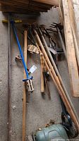 Photo showing a collection of garden tools on concrete floor including hoes, a sickle, and weed puller