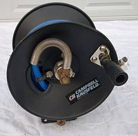 View of the black metal air hose reel with blue hose coiled inside and brass fittings visible. Shows the Campbell Hausfeld label and mounting bracket.