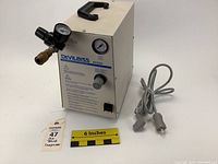 Front angled view of the rectangular beige DeVilbiss 8650D aerosol compressor showing handle, pressure gauges, air outlet, cord, and power switch.