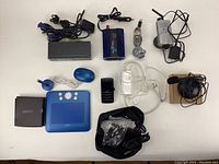 Full view of assorted electronics laid on white surface including Bamboo tablet, video USB splitter, IP camera with antenna, two webcams, power supply cables, blue wireless mouse, and various chargers.