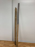 Four hollow metallic CNC guide rails or shafts standing upright, two wrapped partially in brown paper packaging with tape and metal bands, two fully exposed showing shiny but lightly rusted surface. All are approximately one inch in diameter and ranging from 59 to 66.5 inches long.