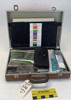 Open briefcase showing a mix of drafting supplies including paper, plastic templates, rulers, and pen cases