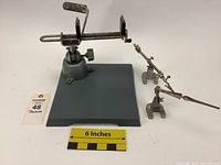 Front view of Panavise vise mounted on gray square base with two separate third-hand jigs to the right and measuring scale showing 6 inches width reference.