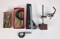 Photo showing three micrometers (two 0-1 inch and one 1-2 inch), Vernier caliper with leather case, and magnetic holder with articulated arm on a magnetic base, all laid out on white surface.