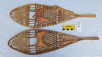 Pair of traditional snowshoes laid flat, showing full wooden frame, rawhide webbing, and attached leather harnesses with buckle straps. Measuring scale and lot tag visible for size reference.