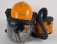 Front angle of the Triton powered respirator showing the orange helmet with clear flip visor, black collar, and the connected orange powered blower unit with hose and filters.