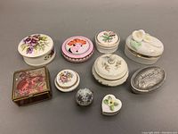Top view showing ten vintage vanity trinket and pill boxes arranged in two rows with various floral, face art, and embossed designs.