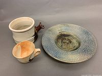 Three pottery pieces displayed: blue-gray textured plate with signature, off-white/orange bowl and mug