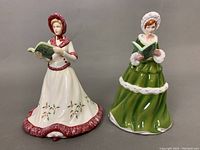 Two Royal Doulton porcelain figurines standing side by side. Left figurine in a white dress with red trim and holly decoration reads a green book. Right figurine in green dress with white fur trim and bonnet also reads a green book.