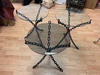 Front side view of three modernist metal chain-link base side tables with round glass tops on wood floor.