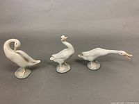 All three white geese figurines shown in a row on a gray surface, illustrating size and pose variations.