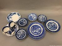 Various Old Willow pattern blue and white plates, small bowls, and teacups with saucers arranged on grey surface.