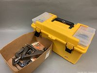 Yellow plastic toolbox with black handle and side compartments next to cardboard box with clamps and pliers inside