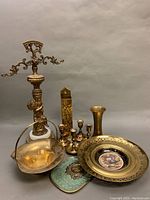 Full lot view showing ornate 22 inch brass cherub piece with a white marble base, brass vase, perforated brass framed porcelain wall plate, candleholders, matchstick holder, small brass container and decorative basket.