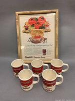 Framed vintage Campbell's Tomato Soup ad with five Campbell's Soup mugs arranged in front showing various soup labels and logos.