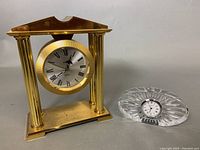 Photo showing the two clocks side by side: a small oval Waterford crystal clock and a larger brass mantle clock with column supports and round clock face.
