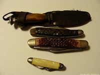 Four vintage knives arranged on a flat surface, various handle materials and sizes, fixed blade knife sheathed.