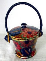 Front angle of antique floral ceramic pot with dark blue wrapped handle and lid knob handle.