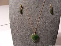 The jade earrings and heart-shaped pendant on gold plated chain displayed on a grey fabric stand.