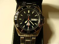 Front view of watch face showing black dial with TFX logo, luminous markers, red second hand, day-date window, and black rotating dive bezel marked 0-60.