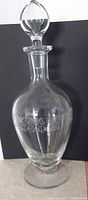 Full view of clear crystal decanter with footed base, etched floral band around body, topped with stopper against black background to highlight design and etching.