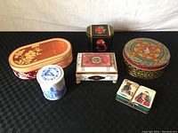 Photo showing seven decorative tin boxes arranged together on a black surface. Includes oval floral tin with lid, white and blue tin, black dragon tin, colorful round tin, pink and white ornate tin with legs, and two halves of a small tin with traditional attire illustrations.