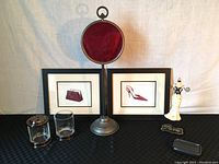 Front view of the lot items including two framed Emily Adams fashion prints (handbag and shoe), glass Q-tip container with lid, glass cup, ceramic cat jewelry holder, ceramic soap dish, and standing copper mirror with red velvet backing.