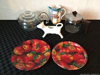 Photo showing entire lot on black checkered surface including two glass coffee pots, German china jug, white cat-shaped item, and two round apple fabric dishes.