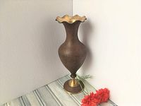 Photo showing full view of brass vase with textured maroon/brown finish and ruffled top edge, positioned on a greyish wood surface with red flowers.