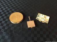 Lot of three items on a checkered black cloth: gold round compact mirror, small square pill box with handle, and rectangular painted pill box.