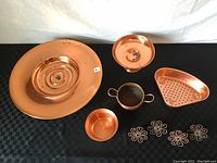 Photo showing all copper items including large rotating cake plate, fruit bowl, small pot, perforated tray, and four napkin rings arranged on black checkered cloth.