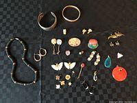 Overview photo showing entire jewelry lot on black checkered background including necklace, bangles, various pins and earrings.