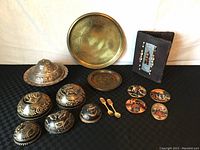 Lot items on black background: etched metal container with lid, brass wall plates, four small hand painted Peruvian art pieces, leather book cover with bead decoration, and gold plated cutlery.