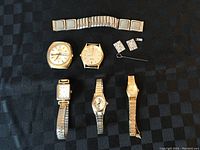 Overview of six watches including men's and women's styles, metal bracelets, and two loose watch links with a small key or tool, arranged on black fabric.