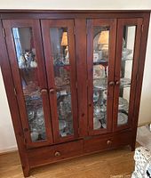 Full front view of the wooden four-door curio cabinet with glass panels showing displayed dishes and crystal items inside, plus two drawers at the bottom with wooden round pulls.