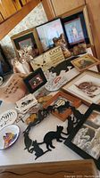 Display of cat-themed trivet, figurines including ceramic cats, carved wooden cat, framed cat prints and wall hangings with cat quotes and designs laid out on table.