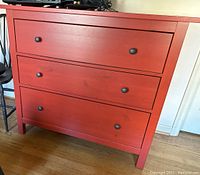Front view of the red chest of drawers showing three drawers with black metal rounded pulls and a sturdy wooden frame.