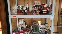 Photo showing two shelves with a collection of Christmas figurines including Santa Claus, snowmen, reindeer, and crocheted angels, along with decorative glass items and ornaments.