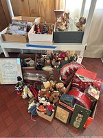 Wide view of the lot showing various boxes containing holiday tins, ornaments, stuffed animals, greeting cards, and figurines arranged on and beneath a small table.