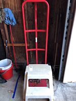 Photo showing red metal hand truck dolly propped against wall with large black pneumatic wheels visible and white plastic step stool resting in front of it on concrete floor.