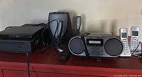 Sony electronics on a shelf including CD/DVD player, video cassette recorder, boombox radio/cassette player, Harmon/Kardon HK 695 speakers and subwoofer, and two AT&T cordless phone handsets with base stations.