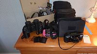 Overview of the vintage electronics lot including the Minolta camera, binoculars, and various camera bags on a wooden surface with cardboard box in background.