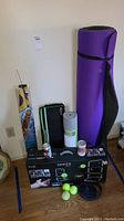 Photo showing full assortment of exercise equipment and related items: purple yoga mat, Gaiam yoga mat, Halex badminton case, 3 tennis balls, kite packaging and The Black Series outdoor ladder game boxed.