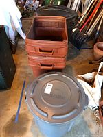 Three brown rectangular plastic garbage bins stacked on top of each other without lids, front view of a dark gray Rubbermaid BRUTE garbage can lid in front