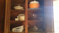 Wooden cabinet interior with shelves holding various glass and ceramic kitchenware items including Glasbake dishes, mixing bowls, and serving bowls.